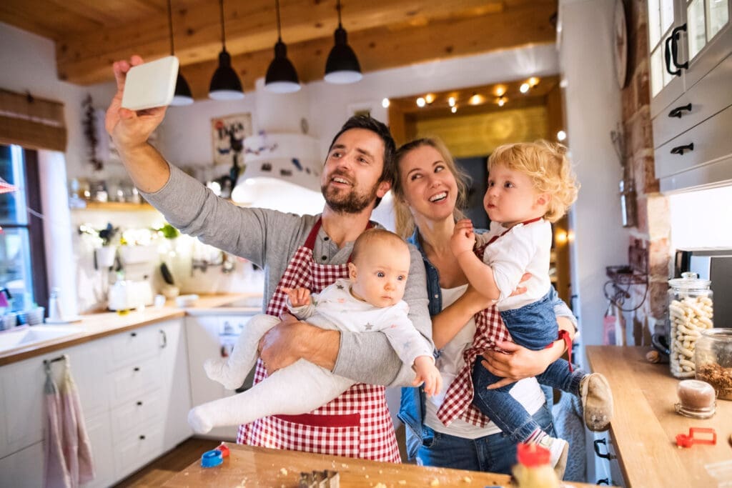 Beautiful young family making cookies at home. Father, mother. toddler boy and baby taking selfie with a smartphone to put in their Modern Baby Book